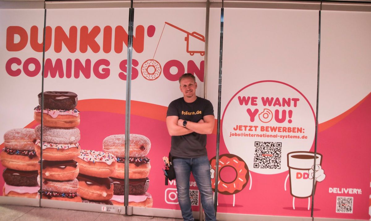 Folierte Bauzaun-Wand mit Dunkin' Coming Soon und Recruiting-Botschaft, davor ein Monteur von folieren.de