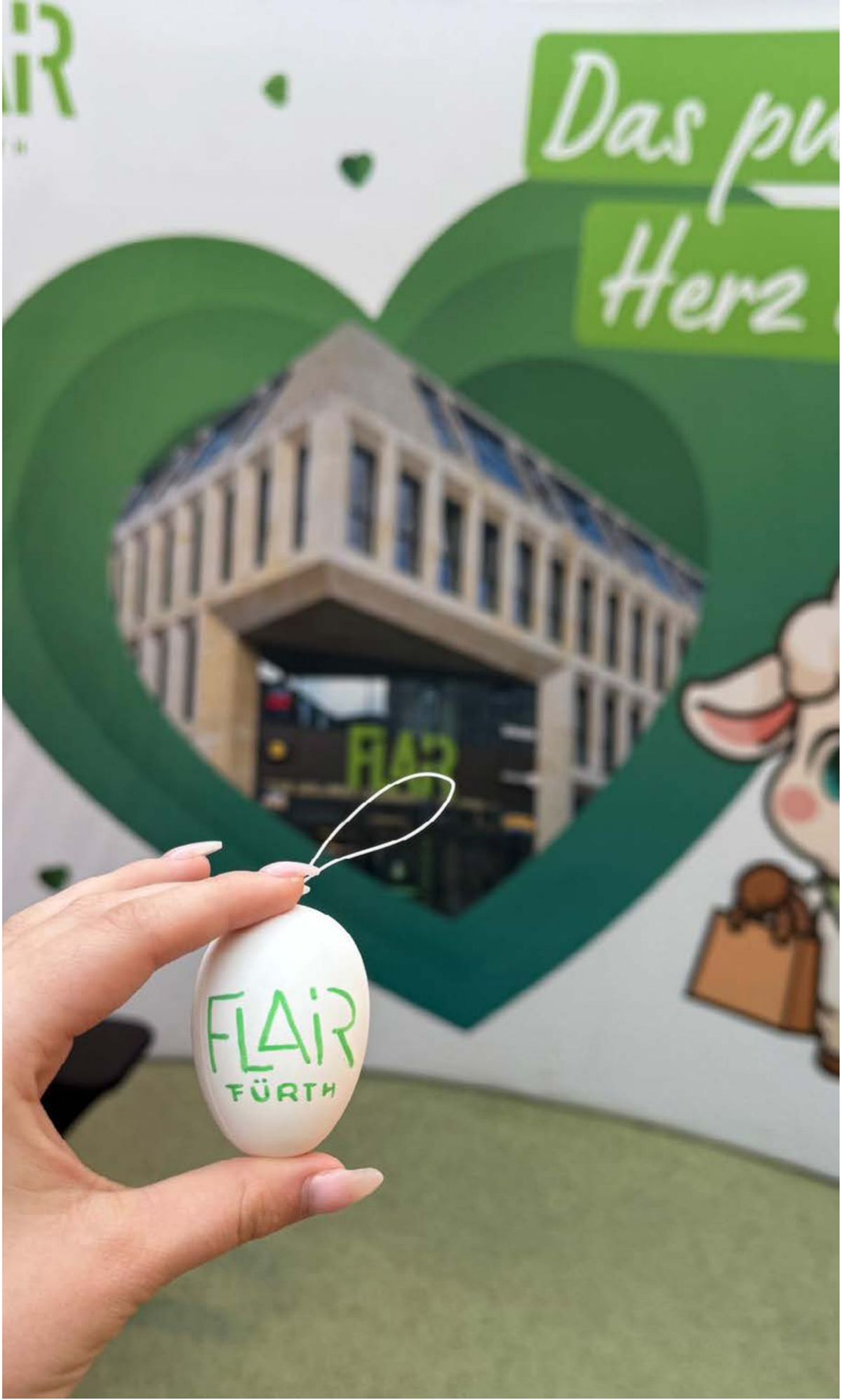 Hand hält ein weißes Osterei mit FLAiR-Logo vor der grünen FLAiRY-Rückwand