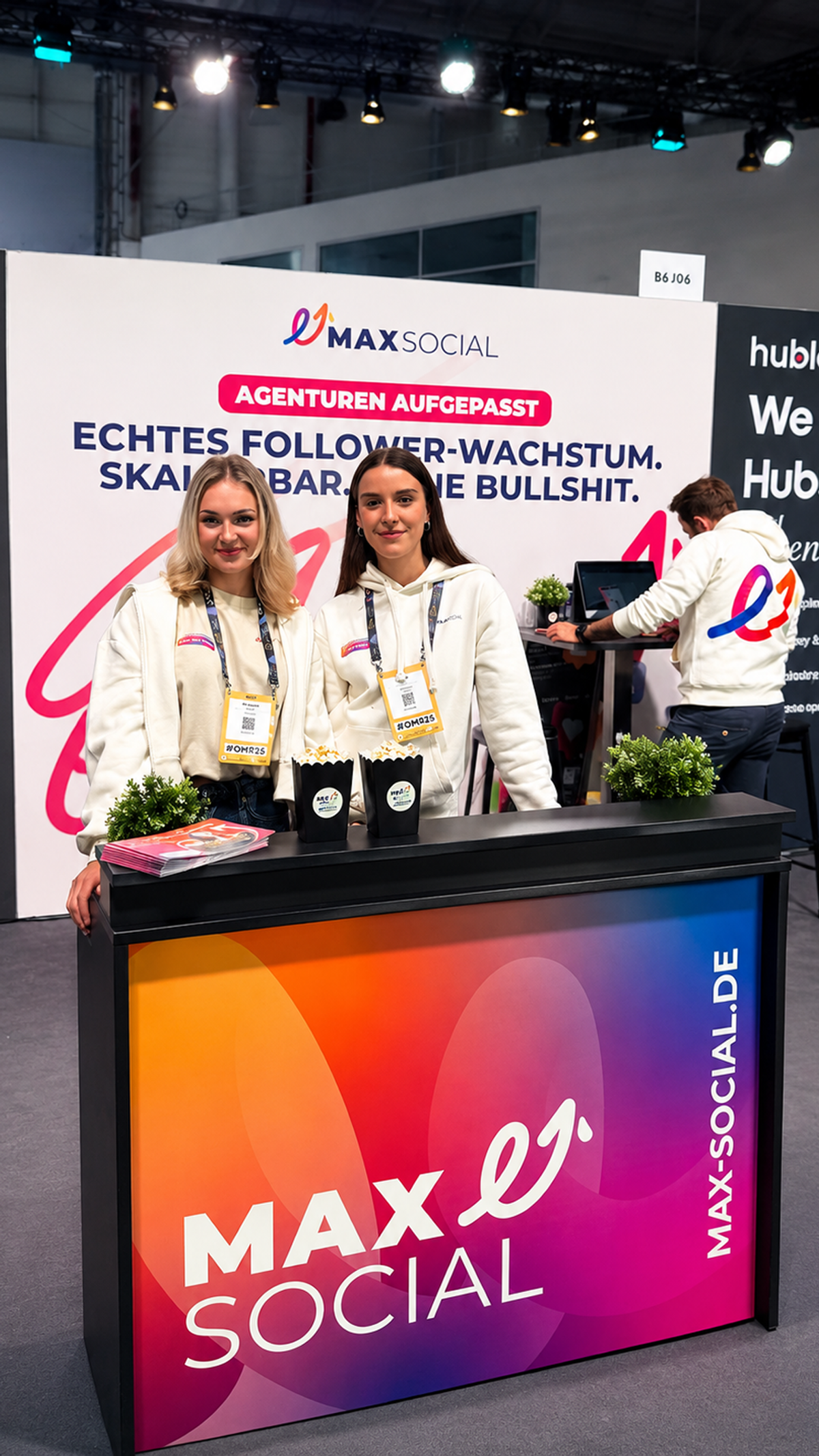 Zwei Mitarbeiterinnen in weißen MAXSOCIAL Hoodies hinter der Theke im Farbverlauf am Messestand B6 J06, dahinter die Rückwand mit Claim