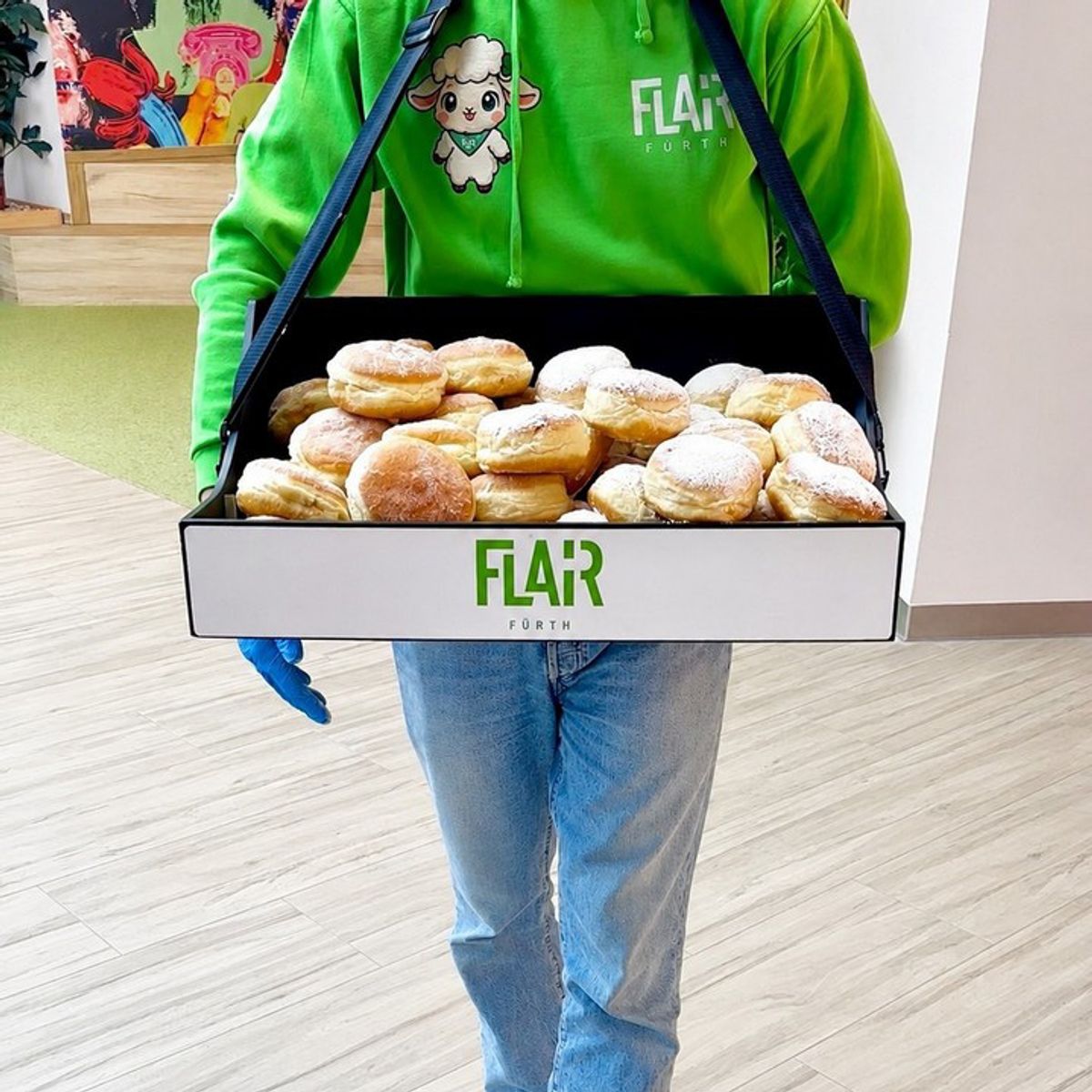 Promoterin im grünen FLAiRY-Hoodie trägt einen Bauchladen mit Krapfen und FLAiR-Logo durchs Center