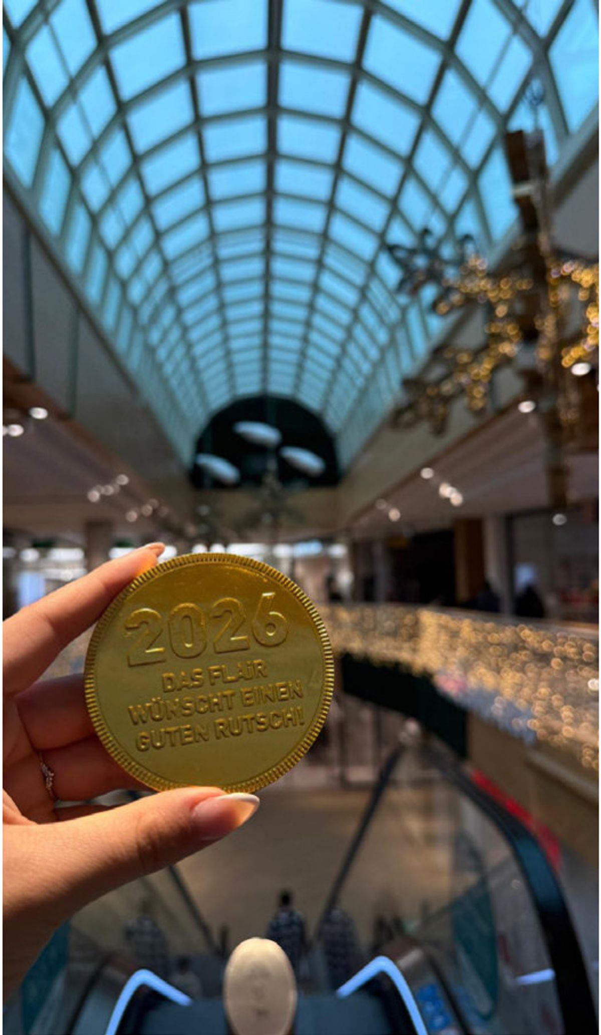 Hand hält einen goldenen Glücks-Schokotaler unter dem Glasdach des FLAiR Fürth