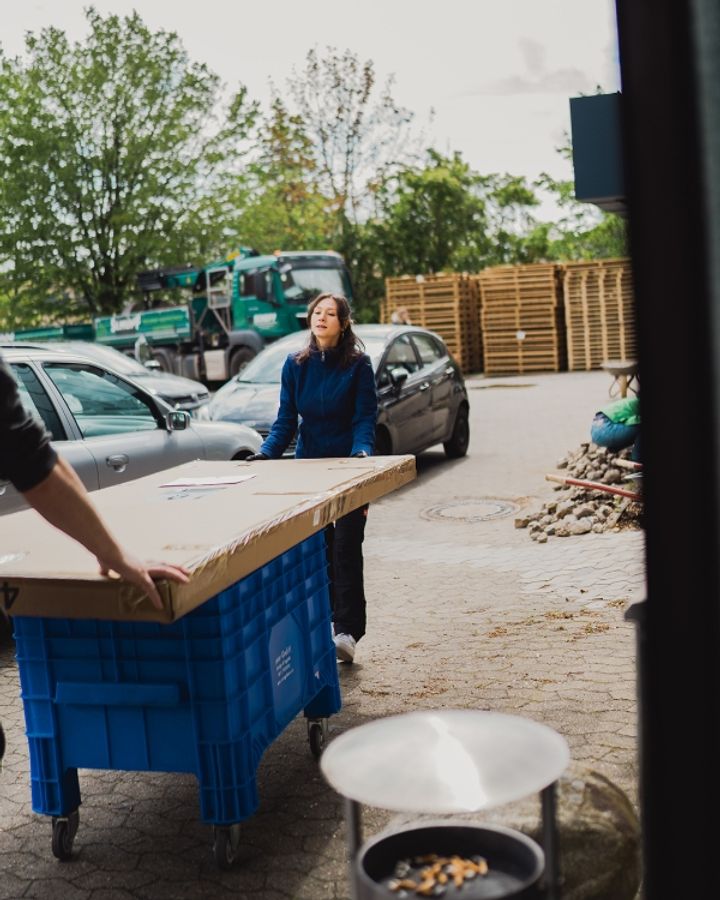 Frau schiebt eine blaue Rollbox mit großem Karton über den gepflasterten Firmenhof, im Hintergrund Paletten und ein Lkw
