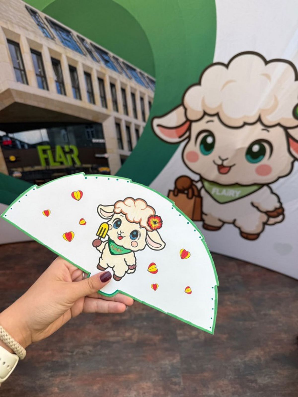 Hand hält einen selbst gestalteten Papierfächer mit FLAiRY-Motiv vor der FLAiRY-Rückwand