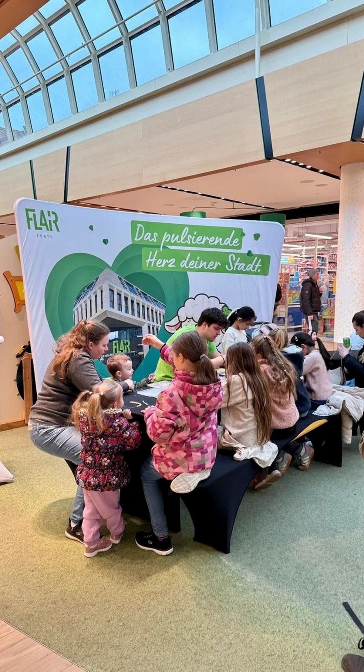 Kinder basteln mit Betreuerinnen an einem Tisch im FLAiR Fürth, dahinter eine Rückwand mit grünem Herz und „Das pulsierende Herz deiner Stadt“