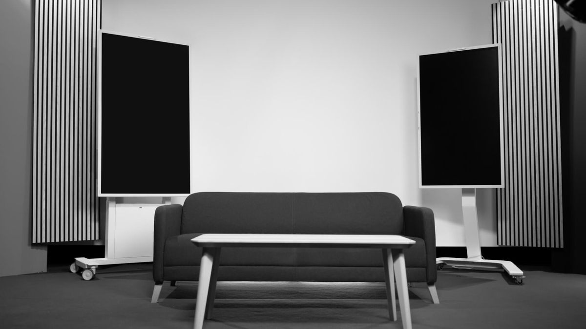 Studioset mit Sofa, Couchtisch und zwei Hochkant-Bildschirmen vor Akustikpaneelen
